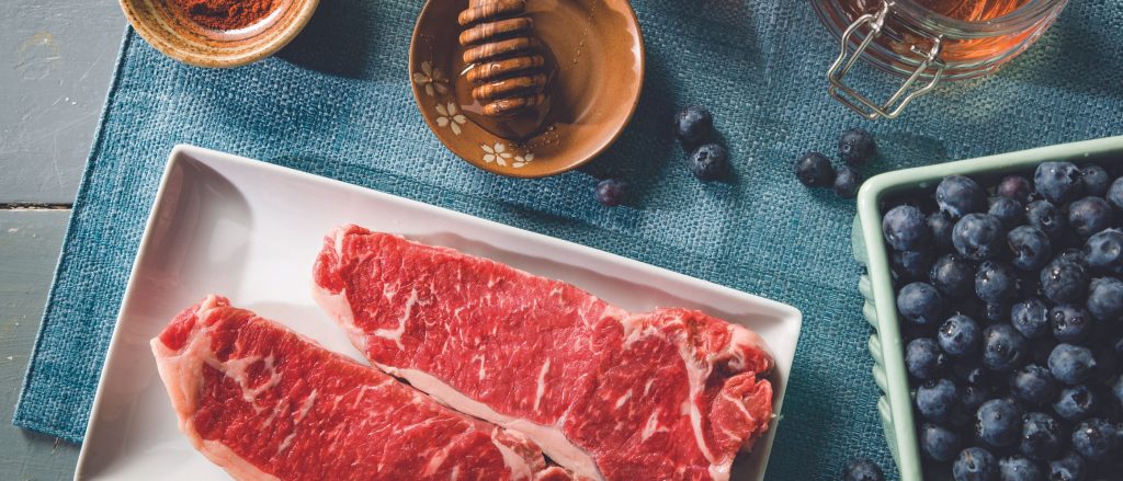 A vibrant jar of Sweet Heat Blueberry Marinade next to fresh blueberries, ready to be used on a delicious cut of Certified Angus Beef ®