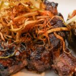 Dry-aged bone-in ribeye steak grilled to medium-rare and topped with smoked cowboy butter and crispy fried leeks, served with a loaded baked potato featuring cheddar cheese, bacon, sour cream, and scallions.