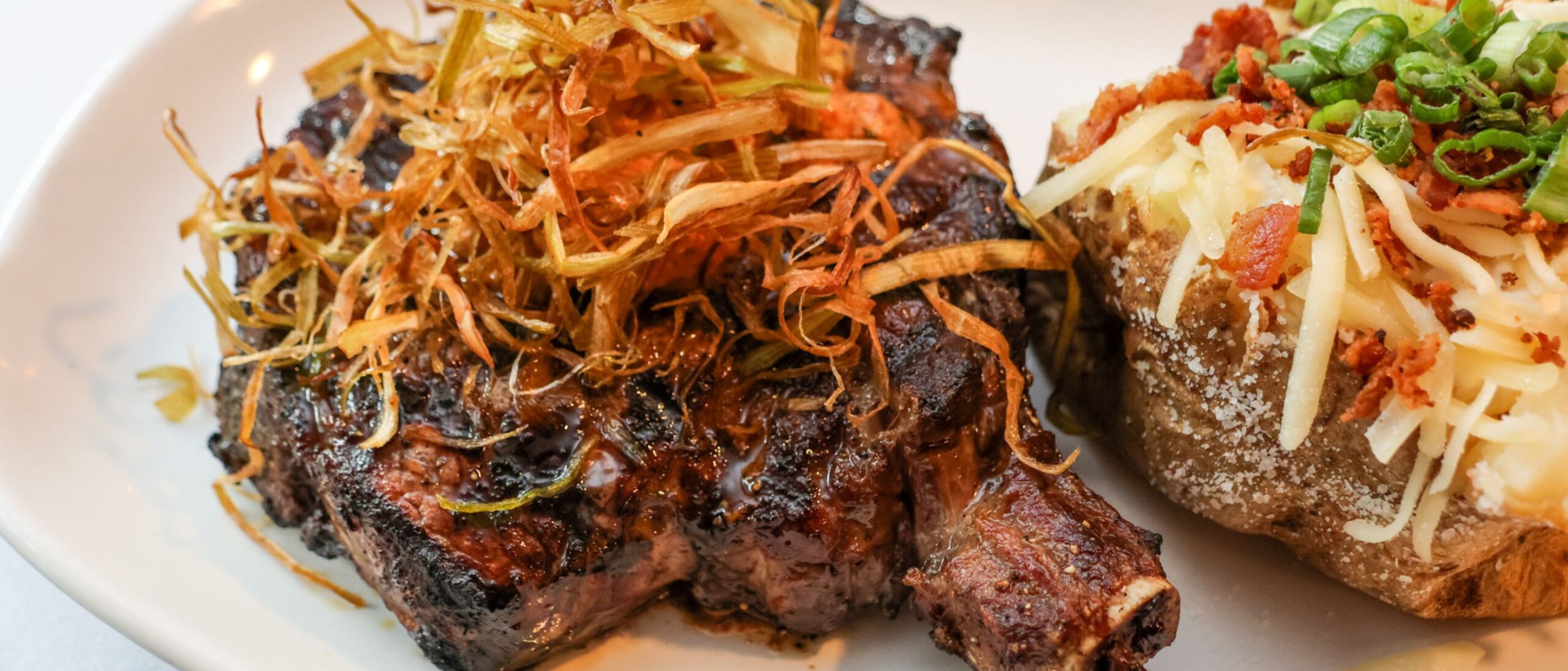 Dry-aged bone-in ribeye steak grilled to medium-rare and topped with smoked cowboy butter and crispy fried leeks, served with a loaded baked potato featuring cheddar cheese, bacon, sour cream, and scallions.
